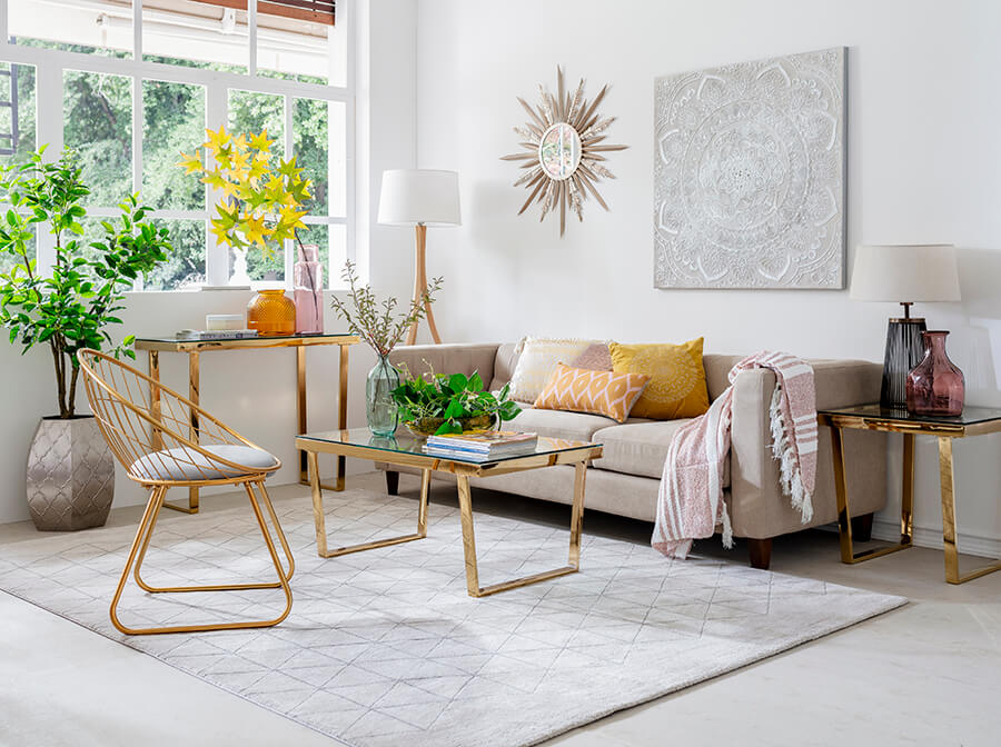 Living y sala de estar con paredes blancas, una gran alfombra blanca y un sofá color arena con cojines amarillos y anaranjado sobre él. A su lado hay una mesa lateral dorada y frente a él, una mesa de centro dorada. 