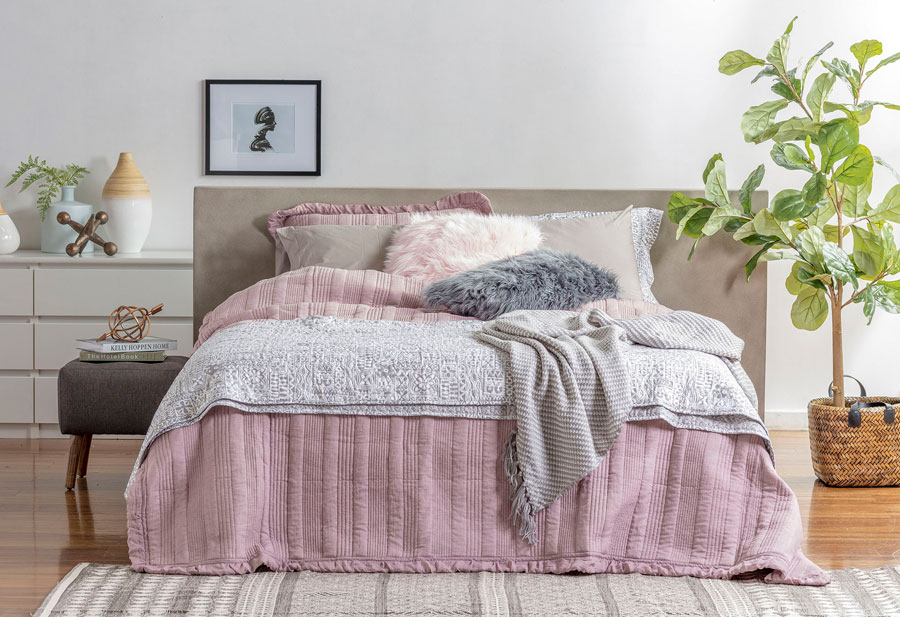 Decoración de dormitorio con ropa de cama de tono gris y rosado. Hay cojines peludos y una manta gris. Al fondo hay una cajonera blanca y, bajo la cama, una alfombra gris.