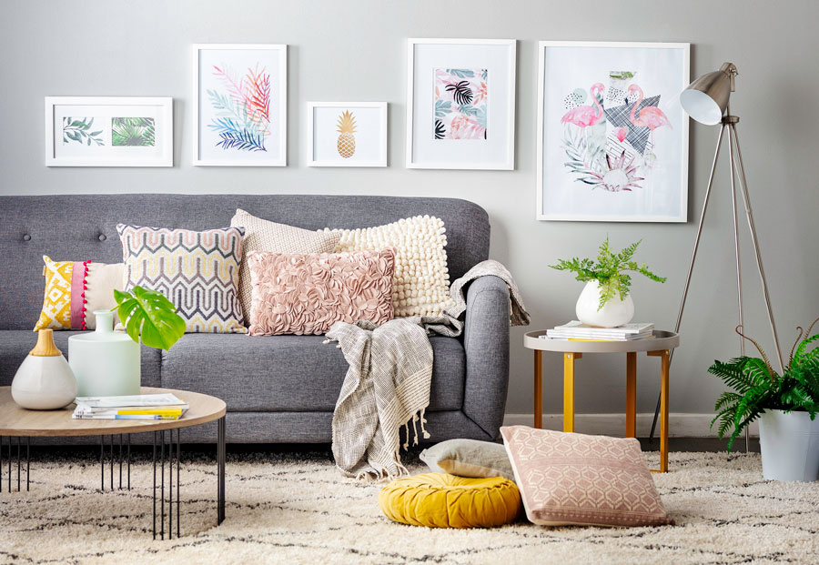 Sala de estar con un sofá gris oscuro con varios cojines multicolor, una alfombra shaggy blanca y una mesa de centro redonda de madera. La pared es gris y hay cinco ilustraciones en marcos blancos.
