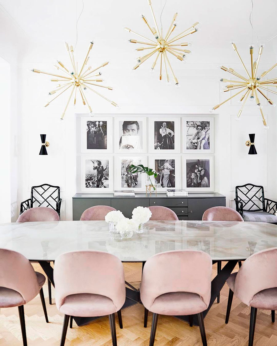 Comedor ovalado de mármol, con ocho sillas rosa velvet con patas negras. Al fondo hay un buffet negro y, sobre ella, una galería muro con ocho marcos blancos con fotografías blanco y negro. A cada lado del arrimo hay una silla negra.