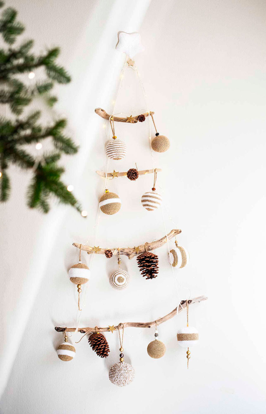 Adorno navideño en forma de árbol, creada con ramas, hilos y esferas de navidad beige y blancos, colgado en la pared.