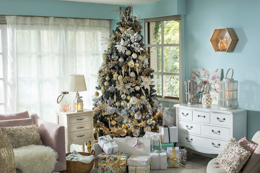 Sala de estar con un árbol navideño alto repleto de adornos dorados y plateados. Debajo del árbol hay regalos y, alrededor, una cajonera, un velador y un sofá y poltrona grises.