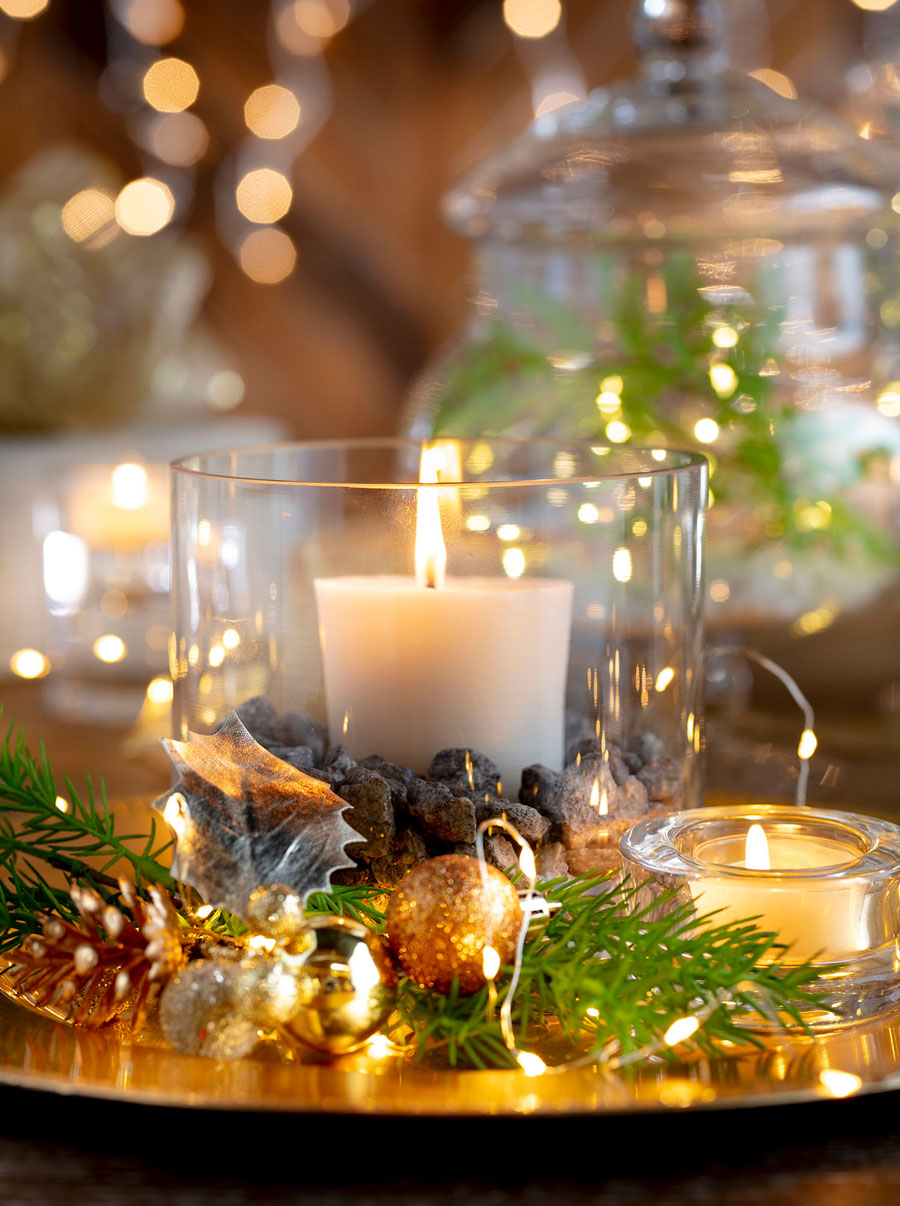 Centro de mesa, una bandeja dorada con una vela al centro. Alrededor de la vela hay otras velas pequeñas, ramitas de pino, guirnaldas de luces y esferas navideñas doradas.