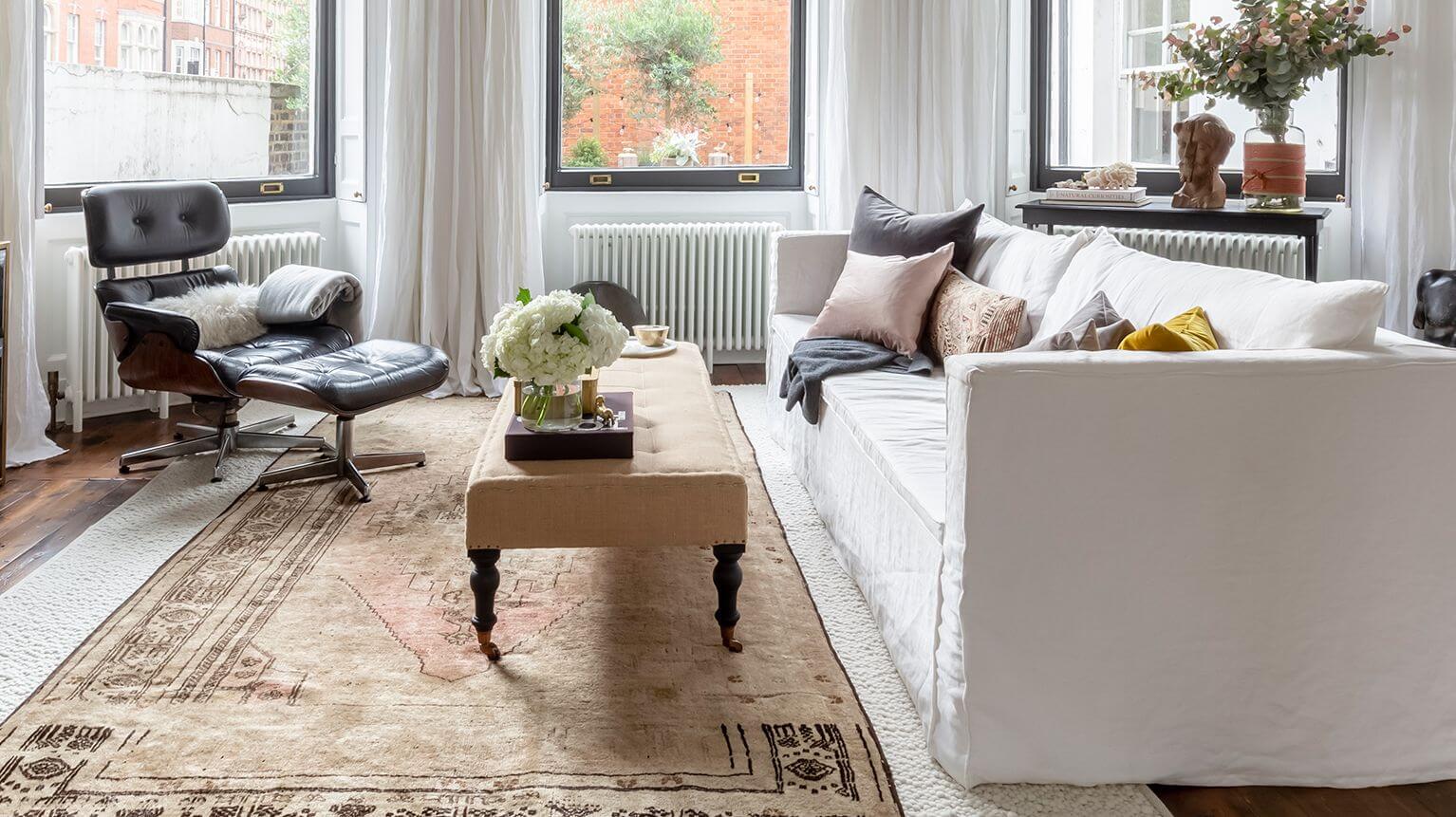 Living con tres ventanas, una alfombra color arena al centro y sobre ella hay un sofá blanco decorado con cojines y mantas, una mesa de centro color beige y una poltrona negra con detalles de madera.