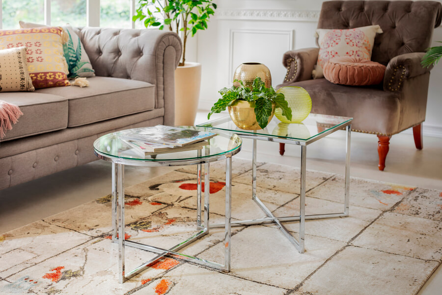 Dos mesas de centro de vidrio con patas de metal, una es redonda y la segunda es cuadrada. Están sobre una alfombra color arena y rodeada por un sofá y poltrona beige.