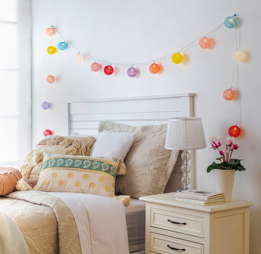 Dormitorio con paredes blancas, cama con frazada y cojines de tonalidades beige y crema y sobre ella una guirnalda de luces de colores, como opción de iluminación interior.