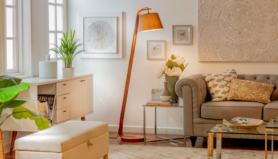 Living con lámpara de pie de madera con pantalla de tela y luz cálida. También hay un sofá gris, una poltrona blanca, una cómoda blanca y varias decoraciones.