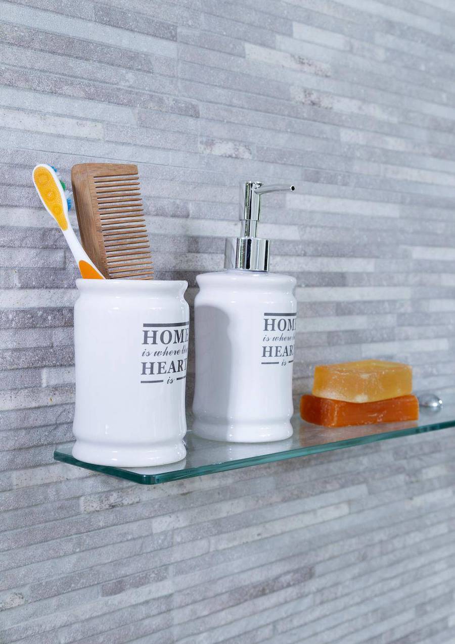 Repisa flotante de vidrio para baño, colgado en una pared de piedra gris. Sobre la repisa hay jabones y vasos de cerámica.