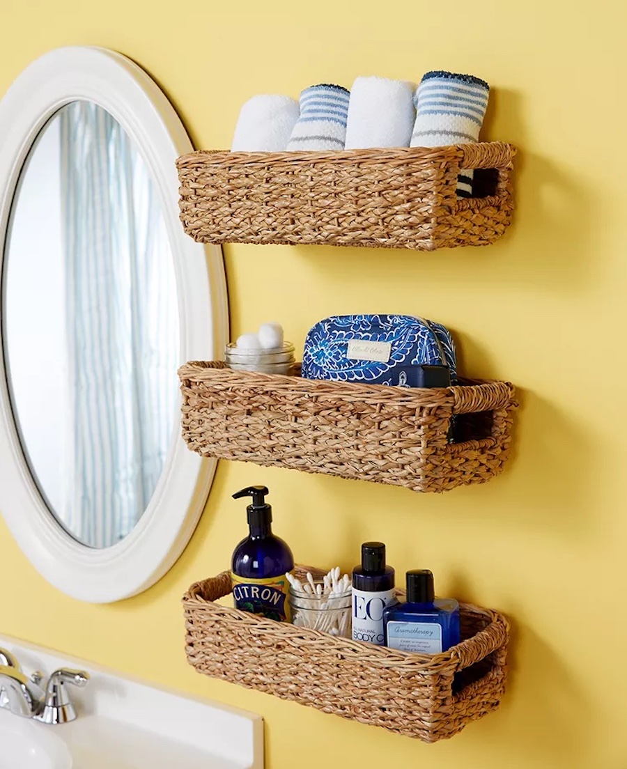 Tres canastos de mimbre colgados en la pared amarilla de un baño, junto a un espejo redondo con marcos blancos. Las canastas contienen productos de higiene personal y toallas.