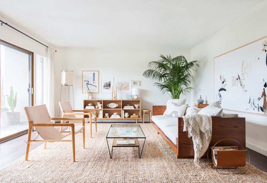 Sala de estar con una gran alfombra color arena y sobre ella hay un sofá de madera con cojines blancos, una mesa de centro y 2 poltronas de madera. 