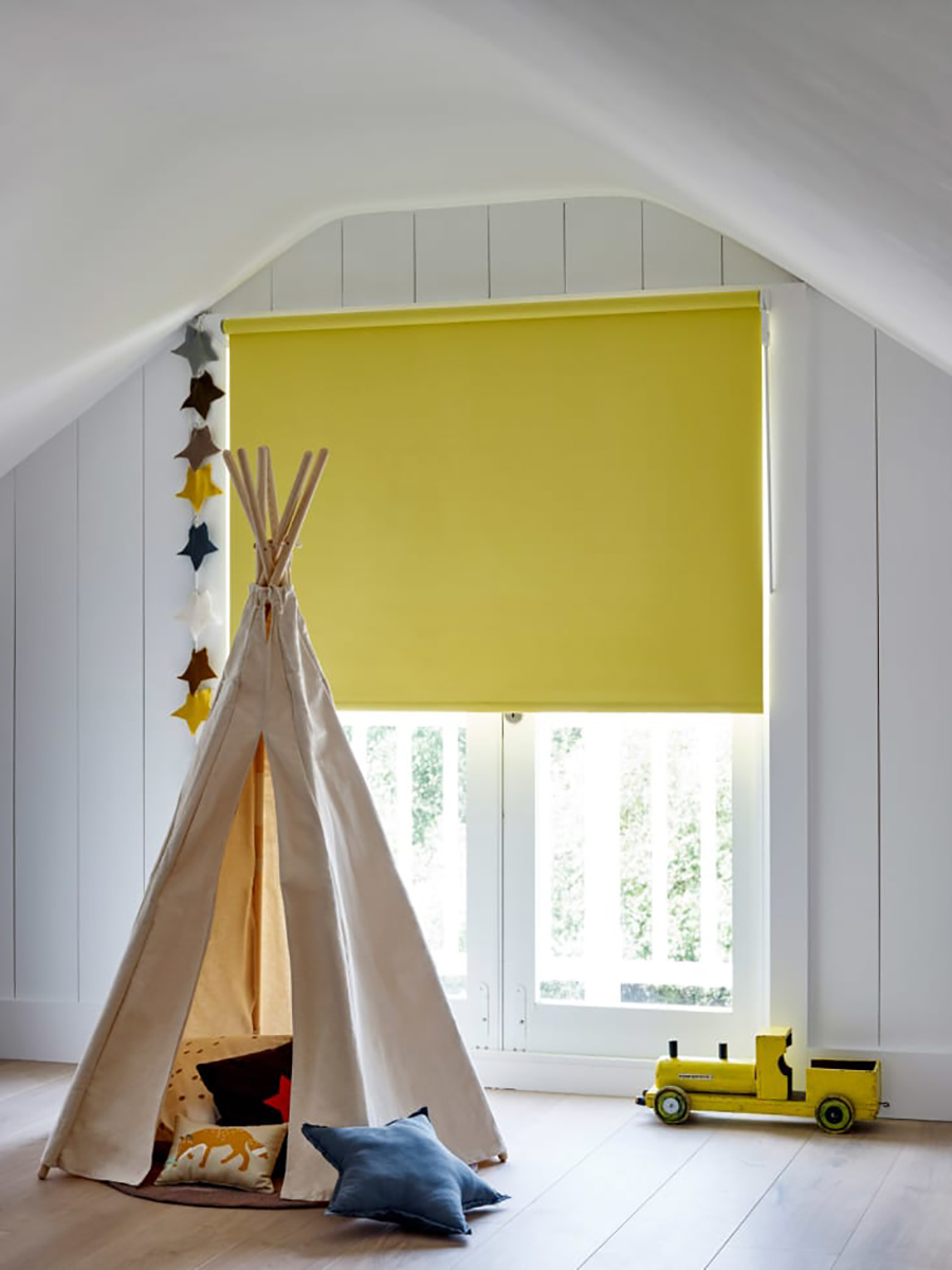 Habitación infantil con paredes blancas y ventana con marcos blancos con una cortina blackout color amarillo. Hay un tipo color crema decorada con cojines con ilustraciones.