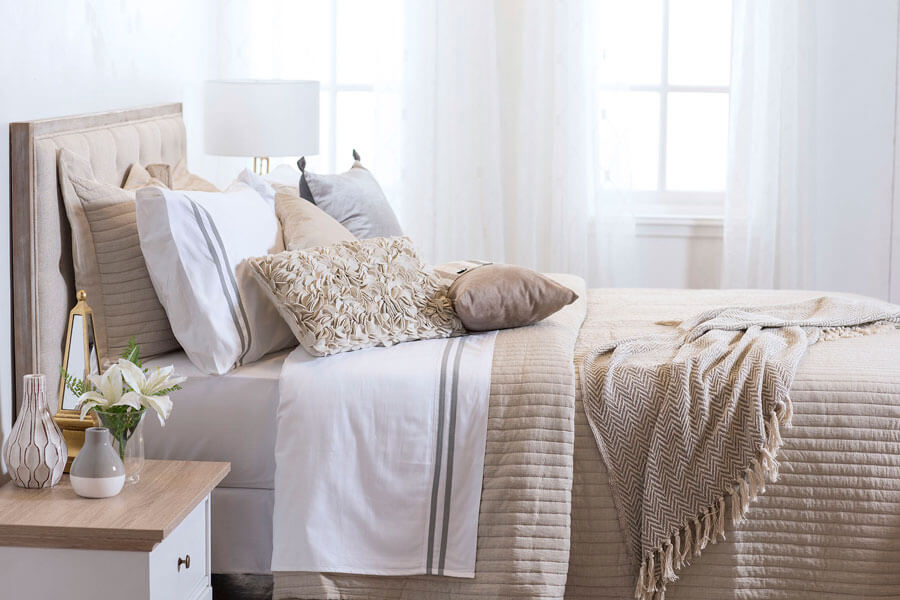 Dormitorio con una cama con sábanas blancas, quilt beige, cojines en color crema y una manta en los mismos tonos. Las cortinas del fondo son blancas.