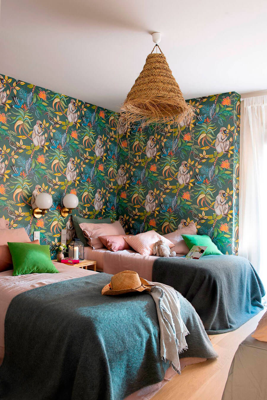 Dormitorio infantil con papel mural con ilustraciones de hojas, monos y plantas. Del techo cuelga una lámpara de fibras naturales. En el lugar también hay 2 camas con sábanas rosadas y cobertor verde oscuro.