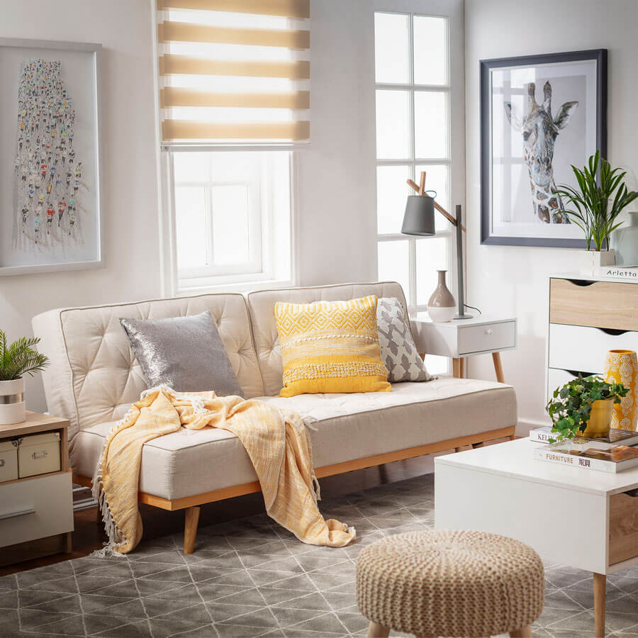 Sala de estar iluminada por una ventana que combina con una cortina roller beige, paredes blancas y un futón blanco con cojines grises y amarillos.