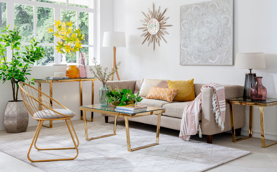 Living de paredes blancas, con un sofá color arena frente a una alfombra blanca que contiene una mesa de centro y poltrona con estructuras de cobre.