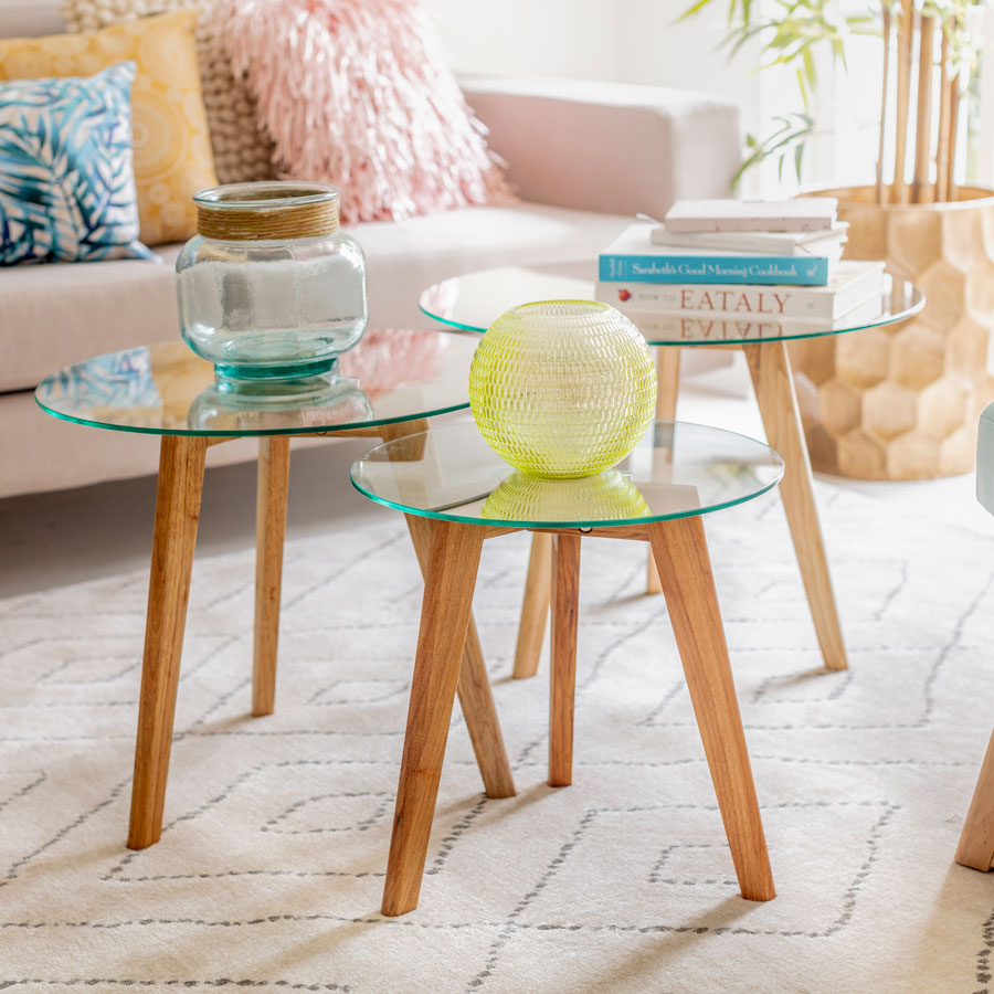 Set de 3 mesas de vidrio de centro redondas con patas de madera en un living. Al fondo se ve un sofá color crema con varios cojines de distintos colores y texturas.