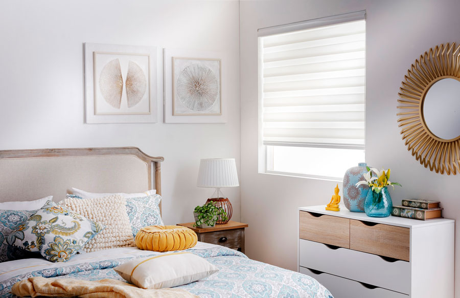 Una habitación con una cama con ropa de cama y cojines con patrones azules, una cajonera blanca y cortina roller blanca.