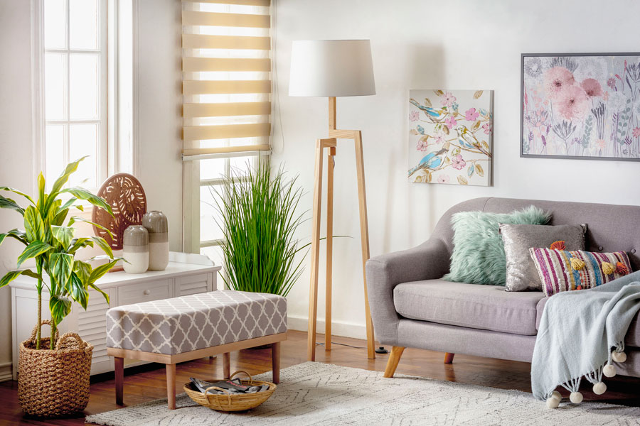 Se ve un living pequeño con cortinas de tela roller dúo color crema, un sofá y banqueta grises, alfombra, lámpara y arrimo blanco.