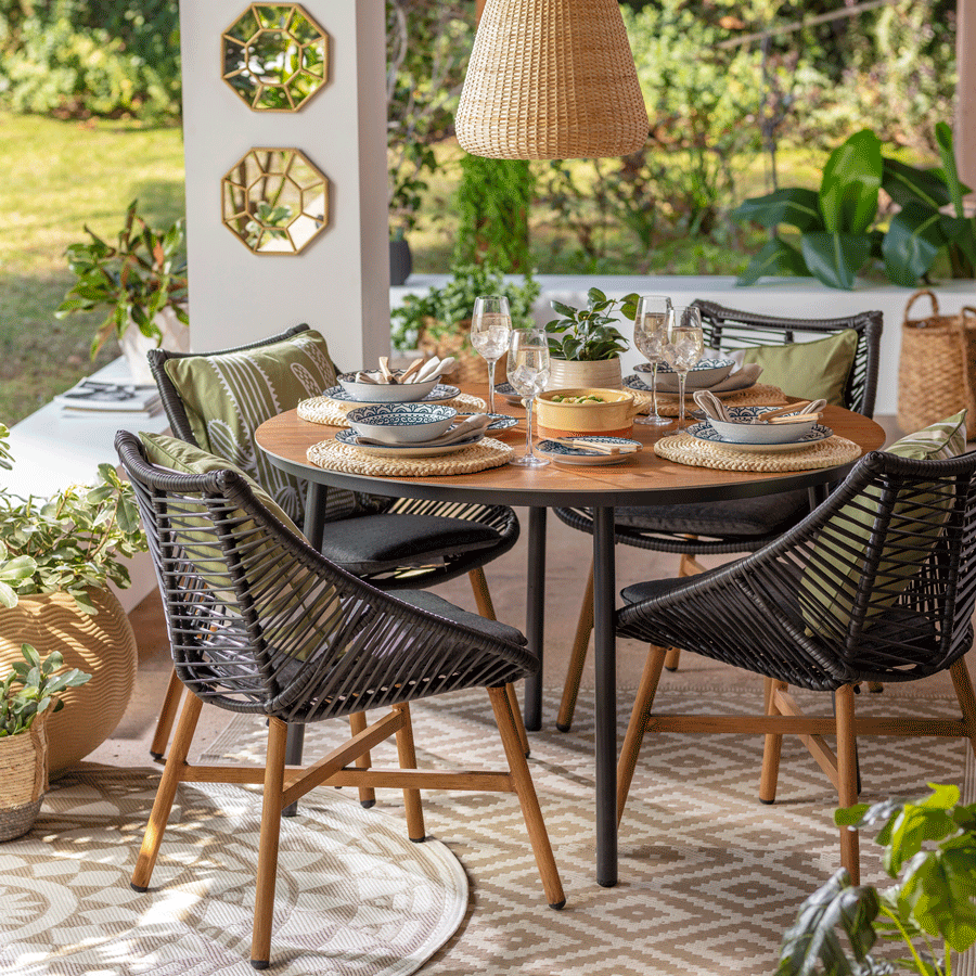 Juego de comedor de terraza redonda con cuatro sillas de madera y detalles negros. Sobre la mesa hay una lámpara colgante de mimbre. Alrededor está decorado con alfombras, espejos y plantas.