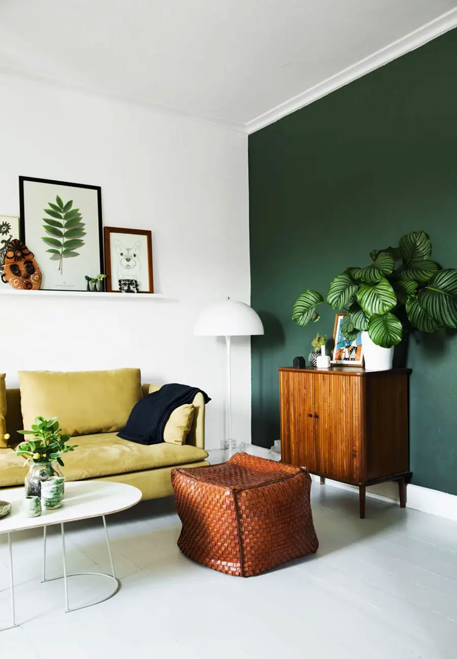 Living con techo, suelo y paredes blancas con solo un pared pintado de verde. Hay un sofá color mostaza, pouf de cuero, mesa de centro blanca y una cómoda de madera que tiene sobre ella una maceta con planta, cuadro y decoraciones.