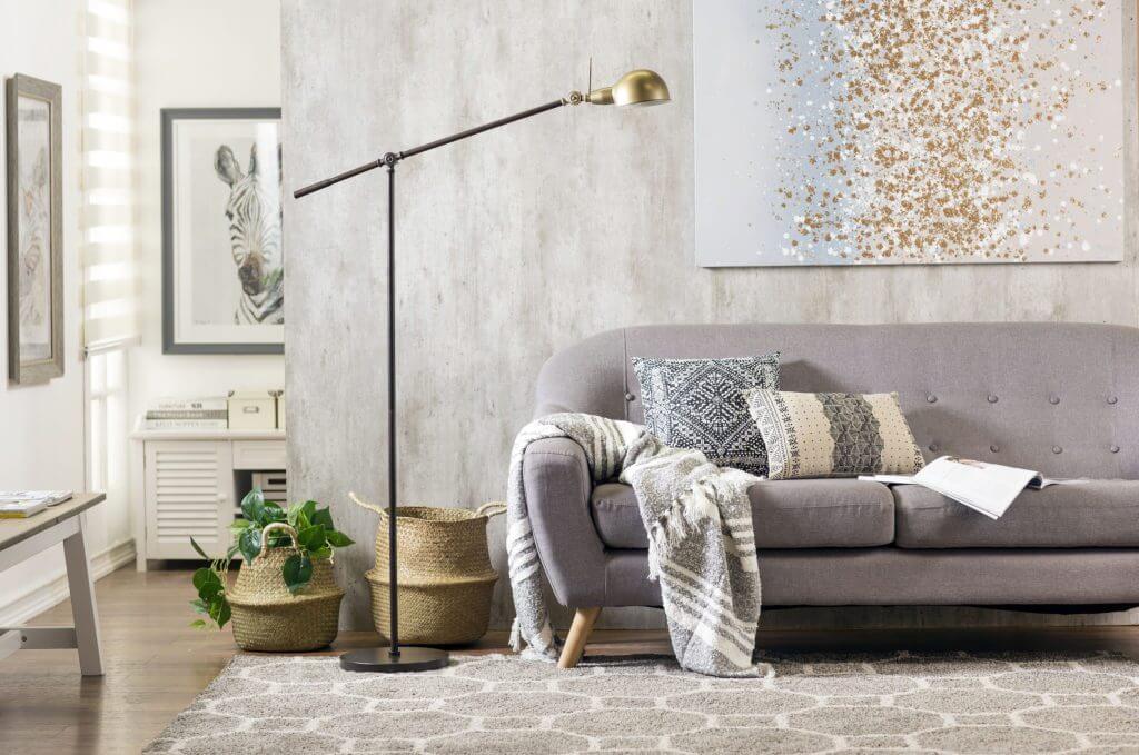 Living de tonos grises con una pared de concreto, sofá gris con cojines y mantas en tonos neutros, alfombra gris con líneas blancas, una lámpara de pie negra con dorado y canastos de fibras naturales.