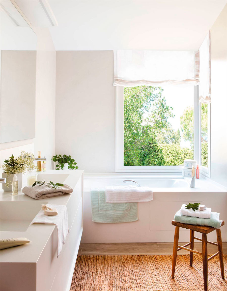 Baño con una tina y lavamanos blanca, piso de madera cubierto por una alfombra naranja y, sobre ella, un piso de madera que sostiene toallas. Al fondo hay una ventana con cortina estor blanca. 