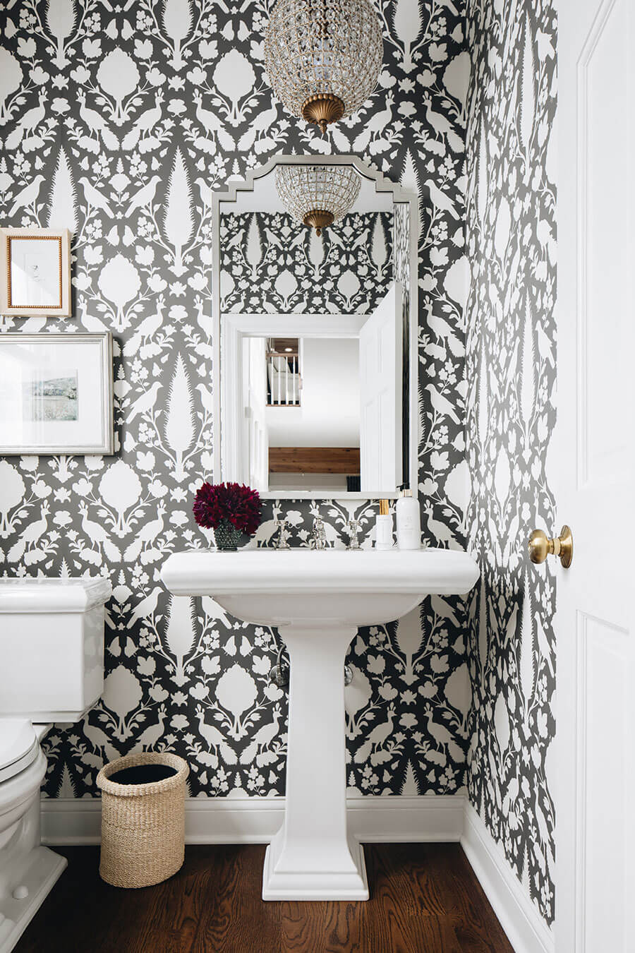 Baño con papel mural con patrones blancos sobre negro estilo vintage. El inodoro, lavamanos y la puerta son blancos. Hay un espejo con marcos plateados y una lámpara colgante con cristales.