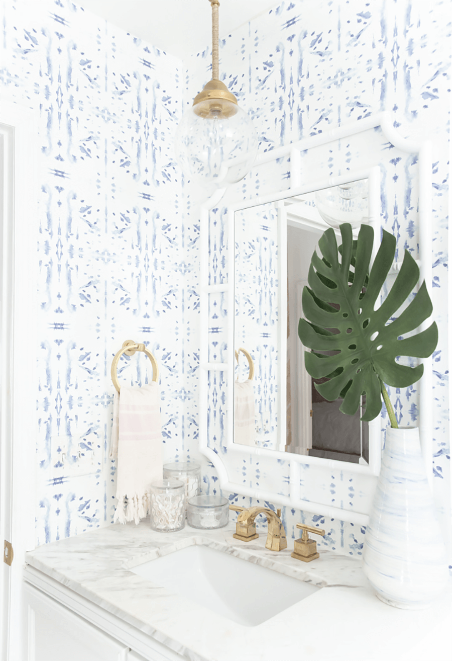 Papel mural para baño con color base blanco y patrones geométricos azules. Hay un mueble lavamanos blanco con cubierta de mármol y grifos dorados, un espejo con marcos blancos y una lámpara colgante dorada con pantalla transparente.