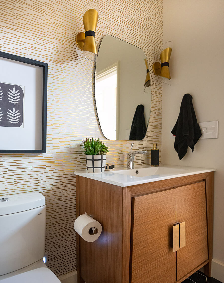Papel mural para baño con base blanca y patrones en línea doradas. El mueble del lavamanos es de madera con cubierta blanca, hay un espejo sin marco acompañado por dos apliqués dorados.
