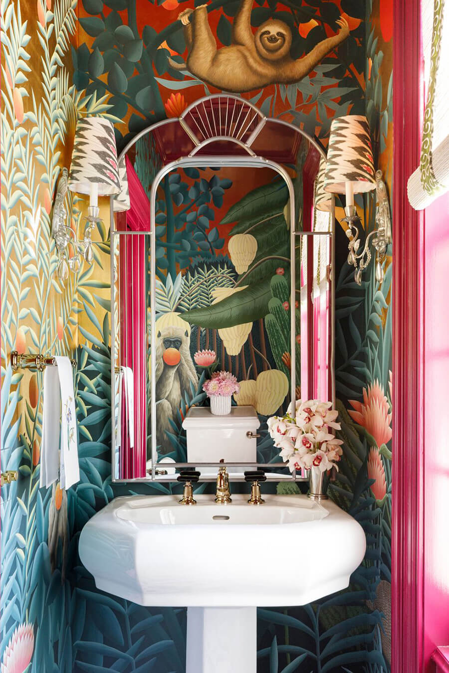 Baño con papel mural con ilustraciones de plantas y animales tropicales. El lavamanos es blanco con detalles dorados y también hay un espejo con marco estilo art deco.