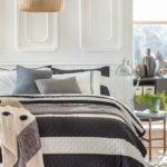 En esta imagen se muestra una cama de tamaño grande, con un cobertor con rayas blancas y negras y cojines en tonos azules y grises.