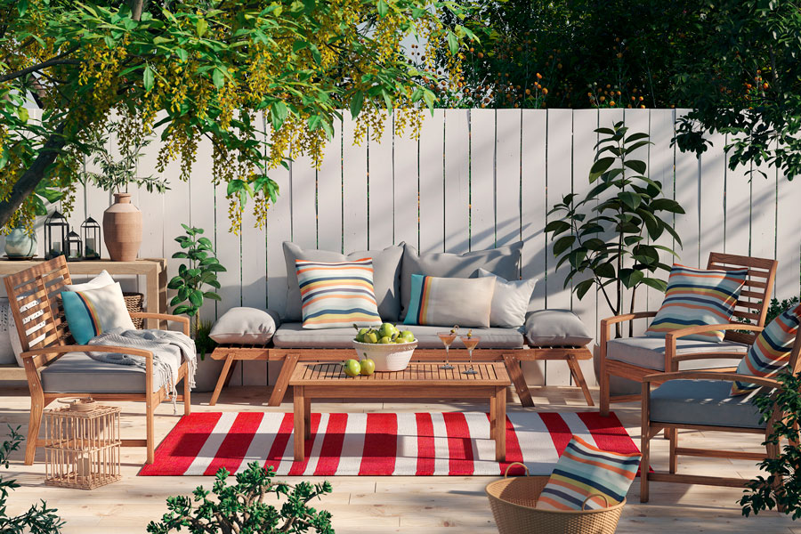 Juego de living para terraza de madera con cojines grises. Están decorados con cojines con listras gris, azul y naranja, y sobre una alfombra con patrón de líneas rojas y grises.