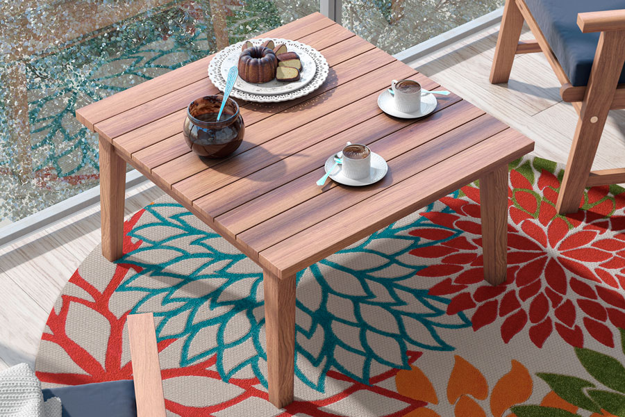 Una imagen de una mesa de madera con dos tazas de café y una tarta. La mesa está situada en un patio con suelo de madera y una alfombra redonda gris con patrones florales multicolor.