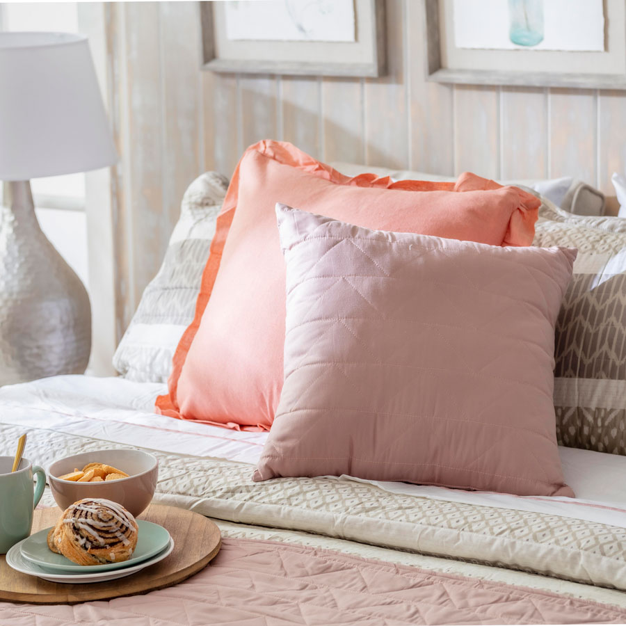 Detalle de una cama con ropa de cama color rosa, crema y rosa palo. Sobre la cama hay una bandeja con platos con un roll de canela y una taza de té