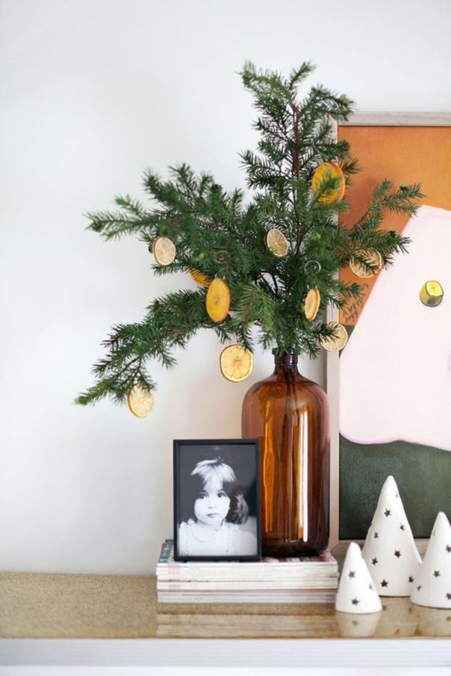 Un botellón de vidrio tiene ramas de pino saliendo de su interior de las que cuelgan adornos navideños hechos con frutas deshidratadas.