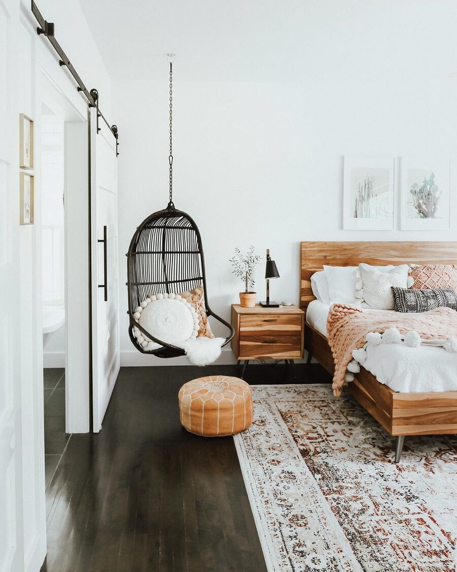 Una silla colgante de fibras naturales negros colgado en un dormitorio con cama y velador de madera, ropa de cama blanca y un pouf de cuero café.