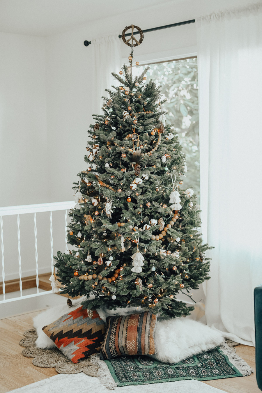 Árbol de navidad decorado con guirnaldas de madera, borlas de macramé y bolas en tonos dorados, en su base tiene alfombras y cojines decorativos estilo boho. 