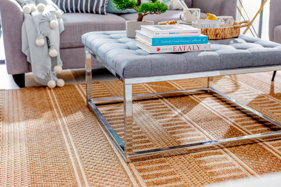 Un living con alfombra de fibras naturales color camello y blanco. Sobre él hay un otomán gris con marcos cromados que funciona como mesa de centro.
