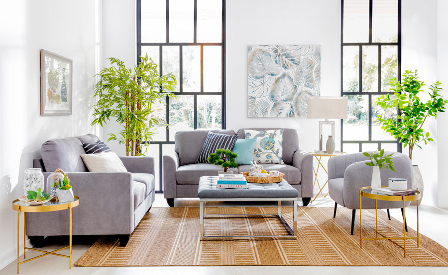 Un living adornado con varias plantas, tres sofás grises, una mesa de centro estilo otomán gris y, debajo, una alfombra de fibras naturales. También hay mesas laterales redondas de vidrio con marcos dorados.