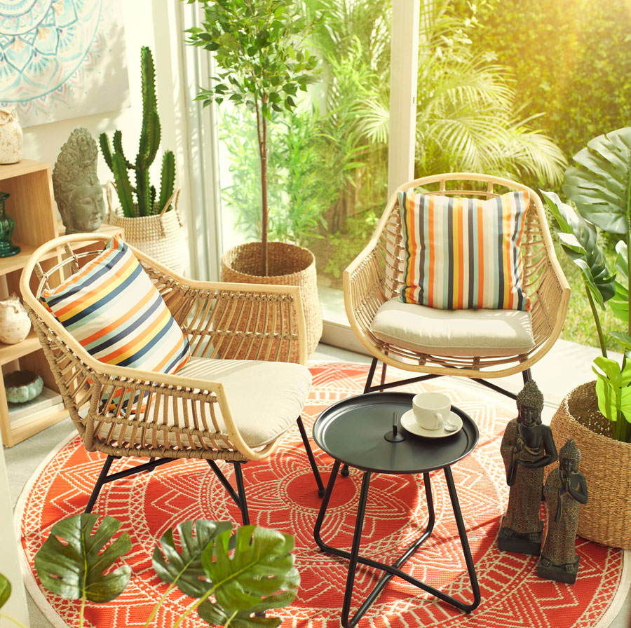 Muebles tipo patio en un living, con un ventanal que conecta con un jardín abierto con plantas tropicales.