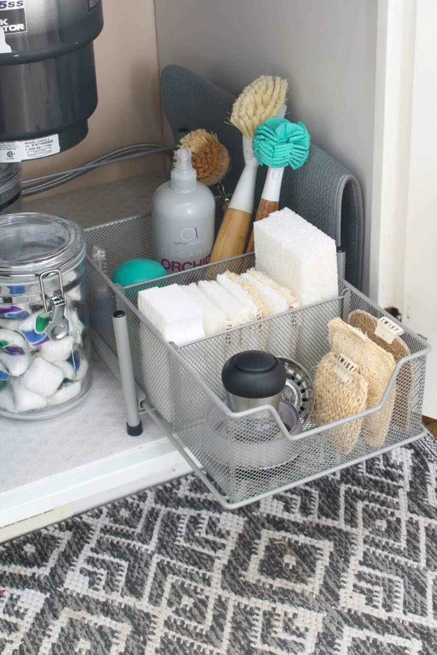 Organizador gris de metal con productos de limpieza junto a un frasco de vidrio que contiene cápsulas de detergente.