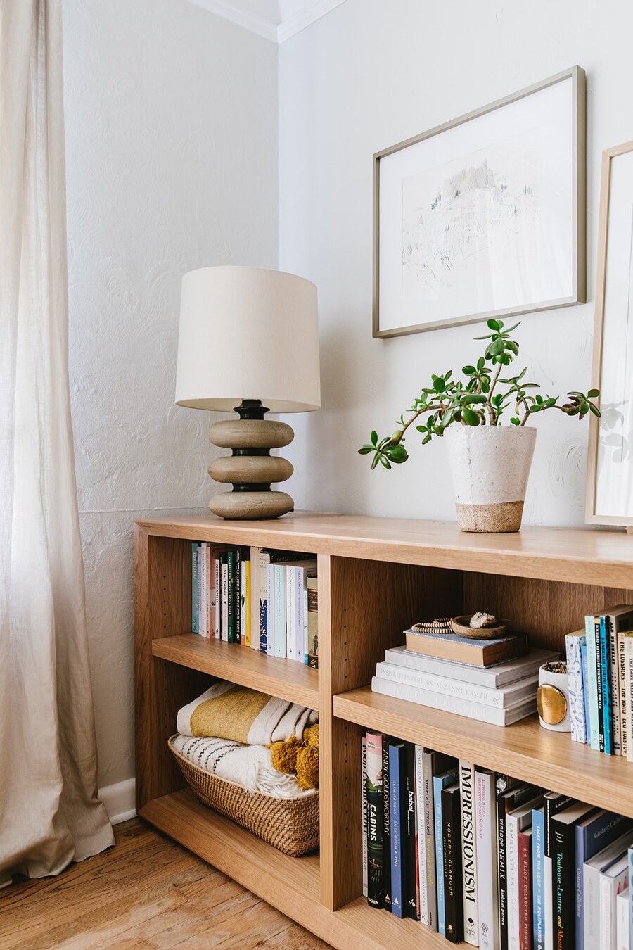Una estantería baja de madera con libros y adornos, como una canasta con textiles, una lámpara y una planta.