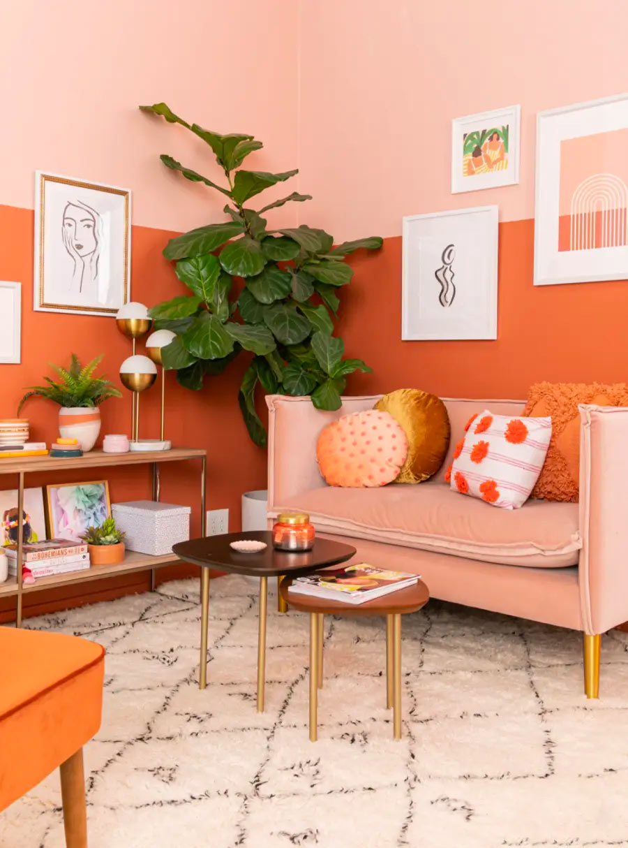 Living con la parte superior de la pared pintada de rosa y la inferior de naranja, con un sofá rosa con cojines naranjos, dorados y blancos. Hay dos mesas chicas de centro de madera con patas doradas y un arrimo de madera con patas doradas.