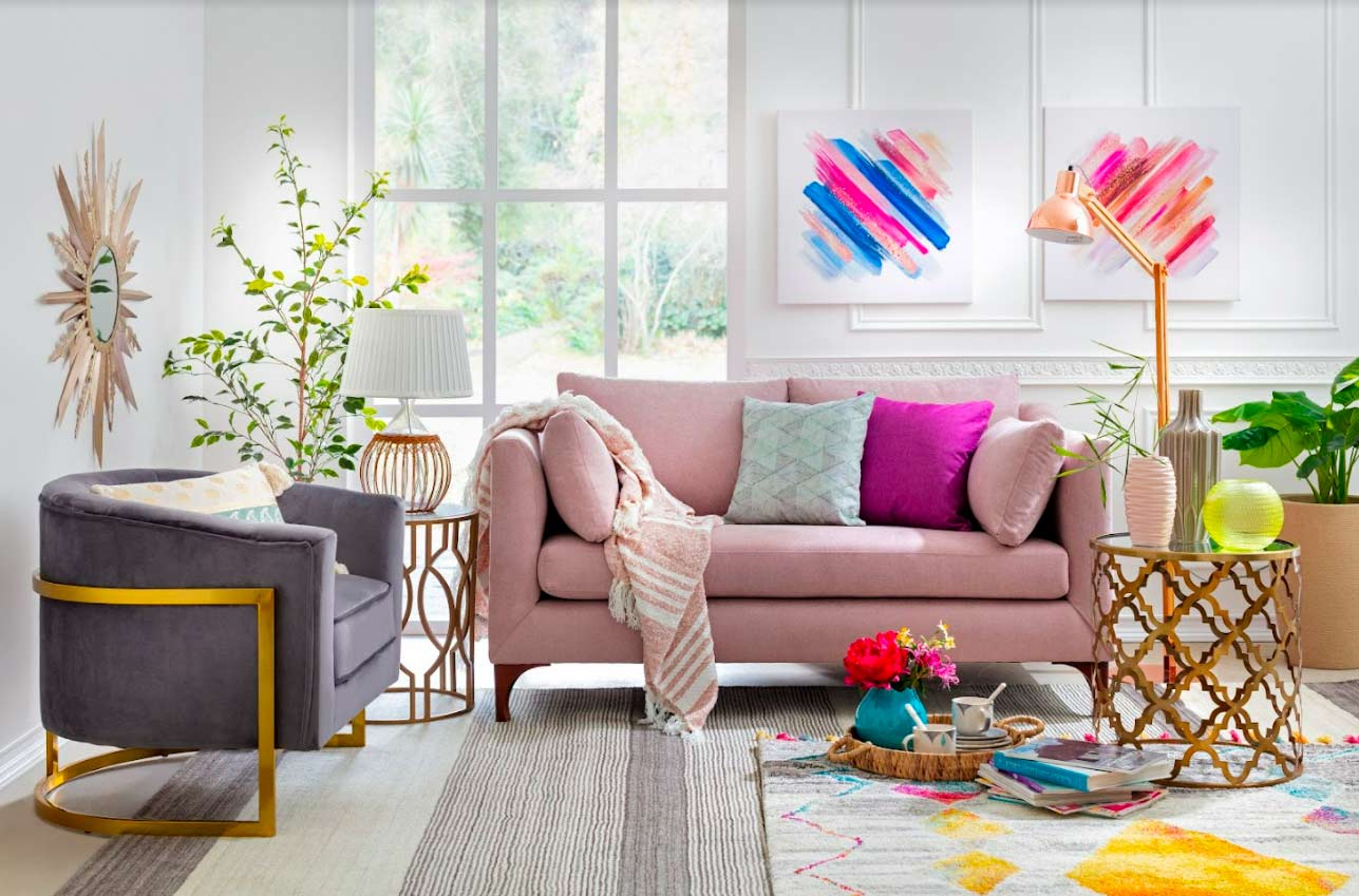 Living con paredes blancas con molduras blancas, un sofá rosado con cojines fucsia, celeste y rosados y una poltrona gris con patas doradas.