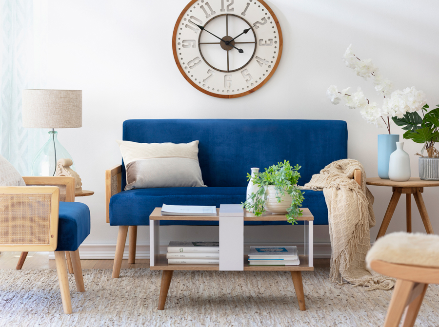 Sala de estar con un sillón azul marino y estructura de madera. Sobre él hay un cojín y manta beige, y desde la pared cuelga un gran reloj análogo. Frente al sofá hay una mesa de centro de dos niveles, de madera clara y detalles blancos. 