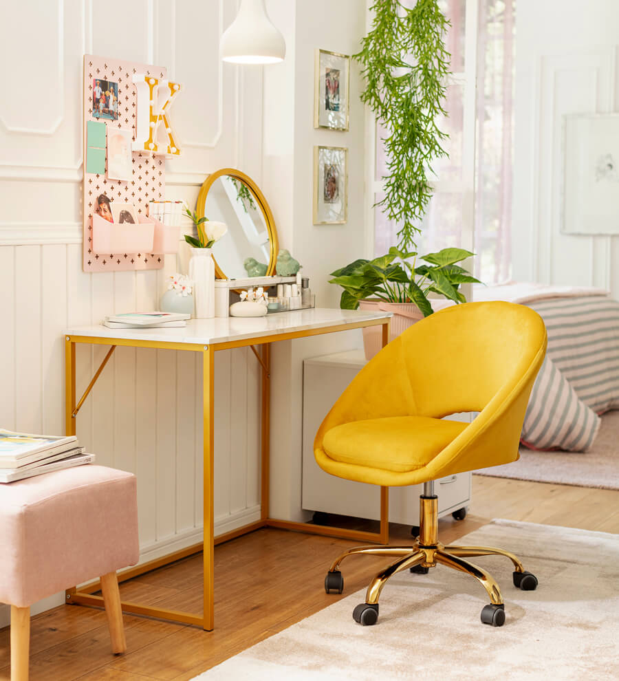 Espacio de trabajo con una mesa blanca y patas amarillas combinado con una silla de oficina de terciopelo amarillo sobre una alfombra blanca. En el escritorio hay jarrones y un espejo redondo con marcos dorados y sobre el escritorio hay un organizador de pared rosa.
