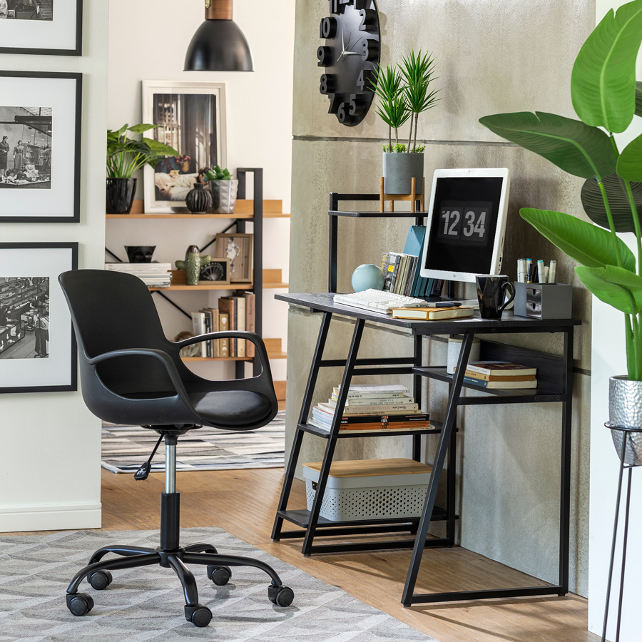 Zona de trabajo o estudio con un escritorio metálico negro, silla de escritorio negra con ruedas y apoyabrazos. Sobre uno de los estantes del escritorio hay una planta, así como también a su costado y en un estante al fondo del pasillo.