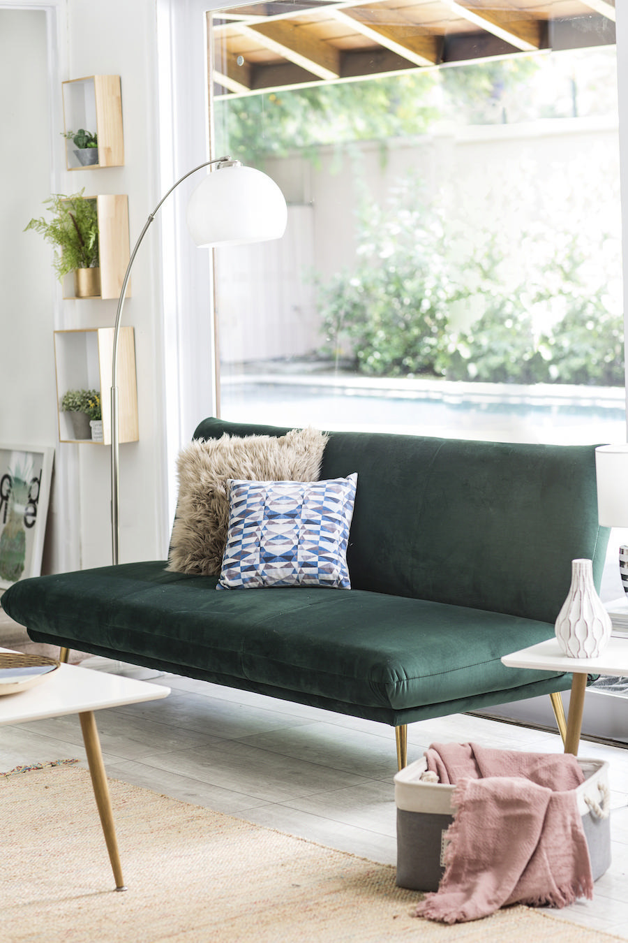Sofá verde petróleo sin apoyabrazos, decorado con un cojín peludo beige y uno estampado blanco y azul. También hay una lámpara de pie, repisas con plantas pequeñas y una ventana detrás del sofá que mira hacia un patio.