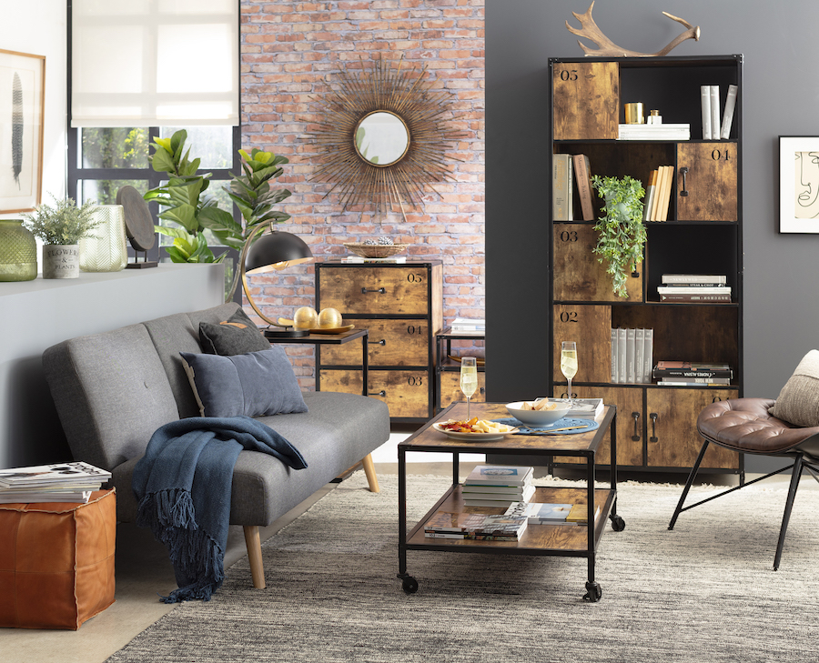 Sala de estar estilo moderno-industrial, con estantes y mesa de centro de madera con bordes negros; sofá gris sin apoyabrazos, pero con cojines y manta en tonos azul marino. También hay plantas que decoran el espacio.