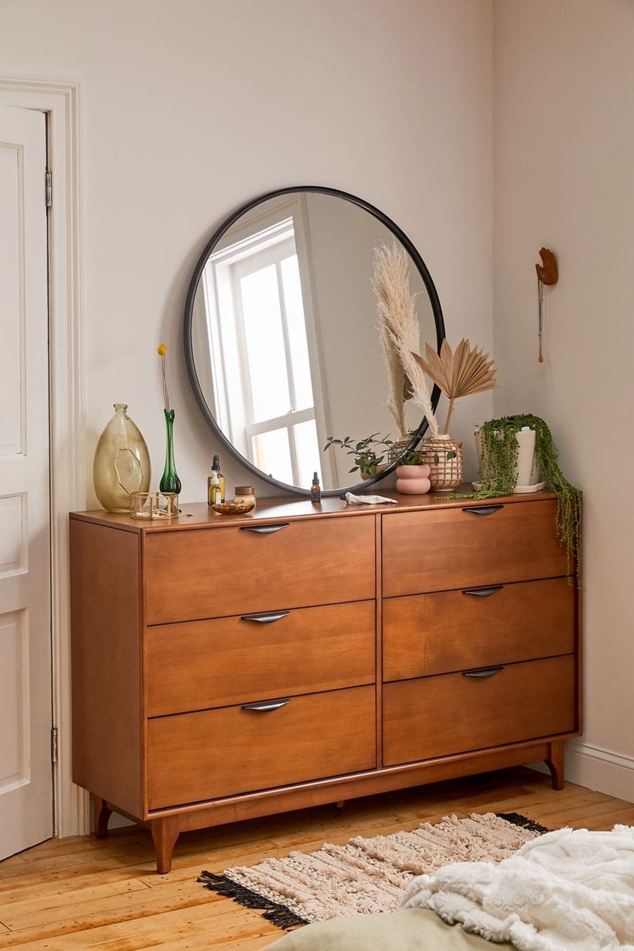 Gran espejo redondo con marco negro sobre una cómoda de madera. Junto al espejo hay frascos, floreros, una planta y otros objetos decorativos. 